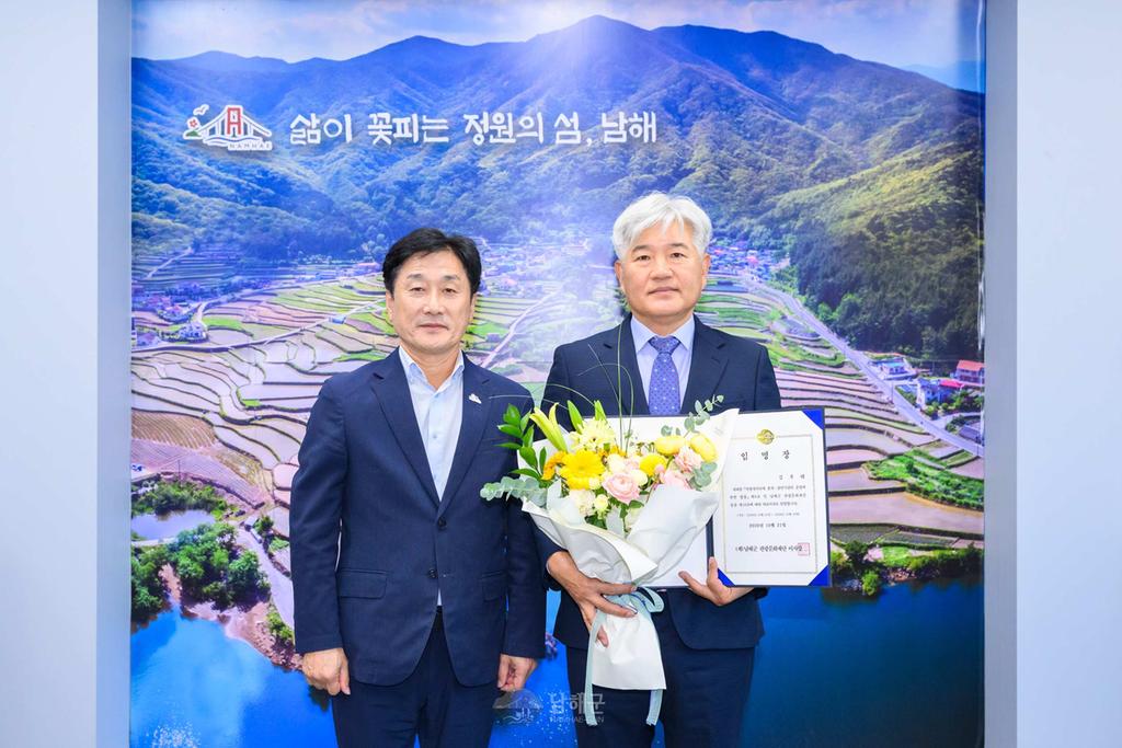 남해의 다랭이논 풍경과 상단에 ‘삶이 꽃피는 정원의 섬, 남해’라는 문구가 담긴 포토월 앞에서, 왼쪽의 류경완 남해군수와 오른쪽의 김우태 남해군관광문화재단 신임 대표이사가 나란히 서서 기념촬영을 하고 있다. 류경완 남해군수는 남색 정장을 입고 있으며, 김우태 대표이사는 꽃다발과 임용장을 들고 카메라를 바라보고 있다. 사진촬영자- 남해군 하철환